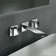 Wand-Waschtischarmatur in Chrom von Ideal Standard mit eckigen Rosetten und einer langen, flachen Auslauf. Montiert an einer grauen Wand über einem weißen Waschbecken.